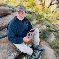 Portrait of Joe Zebrowski: Man wearing a hat, khaki pants and jacket. Sitting on a rock holding a cellphone and sunglasses.